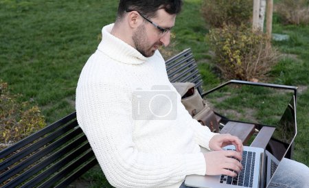 Photo for Young man sitting on bench and using laptop computer in park - Royalty Free Image