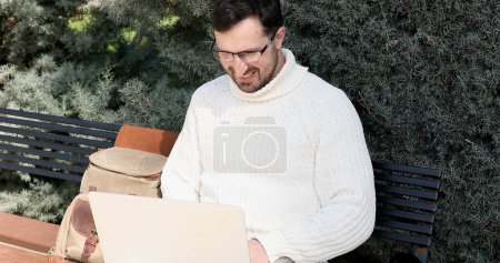 Téléchargez les photos : Jeune homme assis sur le banc et utilisant un ordinateur portable dans le parc - en image libre de droit