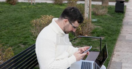 Téléchargez les photos : Jeune homme caucasien assis sur le banc, en utilisant ordinateur portable et smartphone à l'extérieur - en image libre de droit