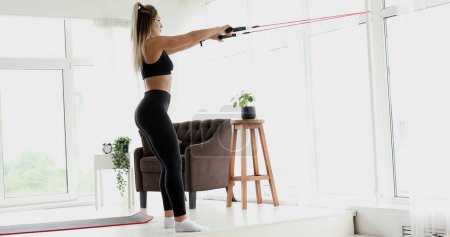 Photo for Sporty young blonde woman during her fitness workout at home with rubber resistance band. - Royalty Free Image