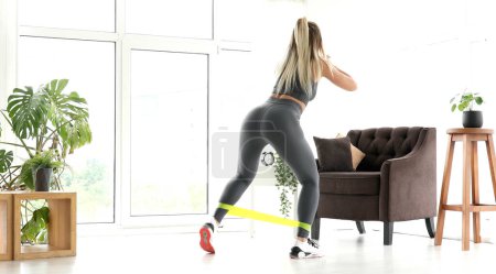 Téléchargez les photos : Jeune femme blonde sportive pendant son entraînement de fitness à la maison avec bande de résistance en caoutchouc. - en image libre de droit