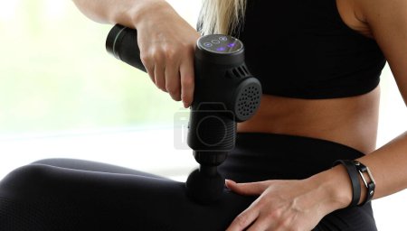 Photo for Young fit woman practicing self-massage technique for muscles applying therapeutic percussive massage gun sitting at home. - Royalty Free Image