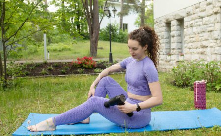 Foto de Vista lateral de una joven mujer en forma sentada en una esterilla de yoga y masajeando músculos con masajeador de percusión al aire libre - Imagen libre de derechos