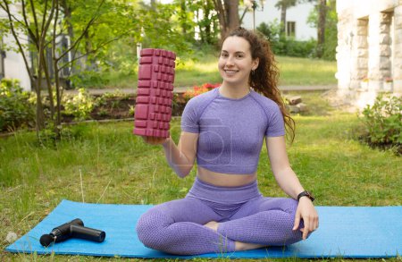 Photo for Young fit woman holding yoga roller and sitting on yoga mat outdoor - Royalty Free Image
