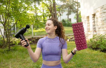 Téléchargez les photos : Jeune femme en forme tenant rouleau de yoga et masseur de percussion en plein air - en image libre de droit