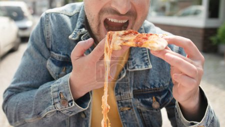 Schöner junger Mann isst draußen auf der Straße ein Stück Pizza.       