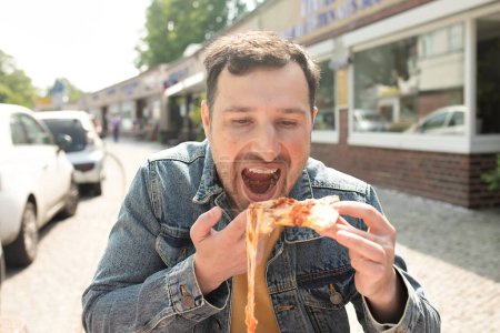 Foto de Joven guapo comiendo una rebanada de pizza afuera en la calle. - Imagen libre de derechos