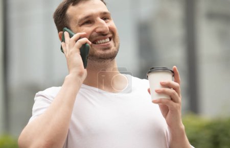 Téléchargez les photos : Jeune homme joyeux boire du café et utiliser le téléphone à l'extérieur. - en image libre de droit