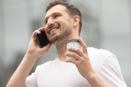 Téléchargez les photos : Jeune homme joyeux boire du café et utiliser le téléphone à l'extérieur. - en image libre de droit