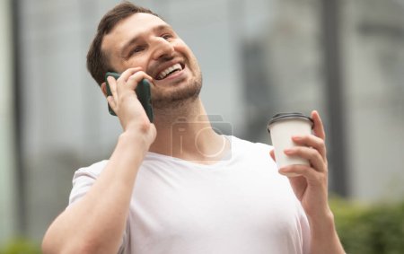 Photo for Young cheerful man drinking coffee and using the phone outdoors. - Royalty Free Image