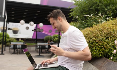 Foto de Joven freelancer sonriendo feliz estudiante satisfecho hombre en camiseta blanca sentarse en el trabajo de banco con ordenador portátil PC descanso relajarse en el parque de la ciudad al aire libre en la naturaleza. - Imagen libre de derechos
