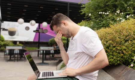 Photo for Young man with headache using laptop, sitting outdoor. - Royalty Free Image