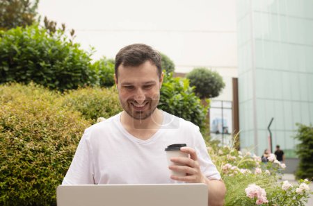 Téléchargez les photos : Beau jeune homme souriant boire du café de tasse en papier et en utilisant un ordinateur portable à l'extérieur - en image libre de droit