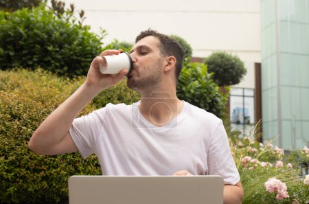 Téléchargez les photos : Beau jeune homme barbu boire du café de tasse en papier et en utilisant un ordinateur portable à l'extérieur - en image libre de droit