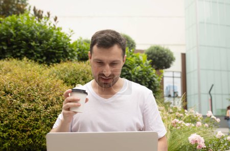 Téléchargez les photos : Beau jeune homme barbu boire du café de tasse en papier et en utilisant un ordinateur portable à l'extérieur - en image libre de droit