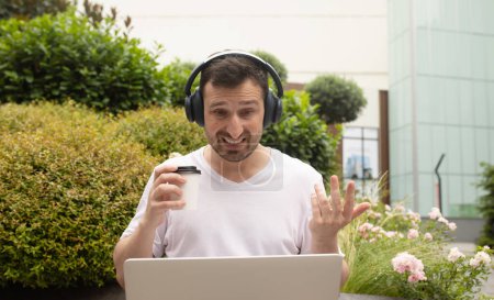Téléchargez les photos : Jeune homme dans les écouteurs en utilisant ordinateur portable à l'extérieur - en image libre de droit