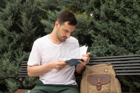 Téléchargez les photos : Jeune homme étudiant portant un t-shirt blanc avec sac à dos lire livre assis sur le banc, se détendre au soleil printemps vert parc de la ville en plein air sur la nature. Éducation concept de lycée - en image libre de droit