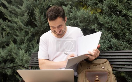Téléchargez les photos : Jeune homme souriant assis sur un banc, en utilisant un ordinateur portable et en écrivant dans un carnet dans le parc - en image libre de droit