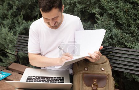 Téléchargez les photos : Jeune homme souriant assis sur un banc, en utilisant un ordinateur portable et en écrivant dans un carnet dans le parc - en image libre de droit