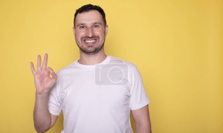 Téléchargez les photos : Bel homme montrant nombre ok symbole, souriant et regardant la caméra sur fond jaune - en image libre de droit