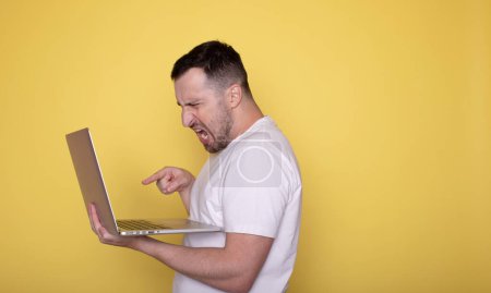 Téléchargez les photos : Jeune homme émotionnel utilisant un ordinateur portable sur un fond jaune. - en image libre de droit
