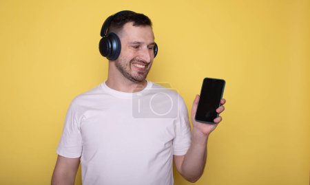 Téléchargez les photos : Heureux homme souriant dans les écouteurs montrant smartphone isolé sur fond jaune - en image libre de droit