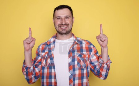 Téléchargez les photos : Jeune homme avec des vêtements décontractés souriant et pointant quelque chose sur fond jaune - en image libre de droit