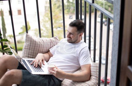 Téléchargez les photos : Bel homme avec ordinateur portable et tasse de café assis sur une chaise sur un balcon. - en image libre de droit
