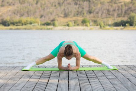 Téléchargez les photos : Femme mince pratiquant le yoga sur la plage près du lac, ayant l'entraînement en plein air, étirant les muscles sur le tapis de remise en forme. Mode de vie actif, soins de santé, concept de soins du corps. - en image libre de droit