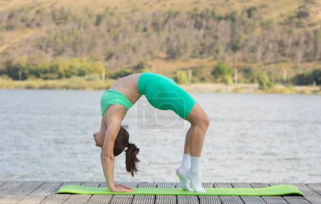 Téléchargez les photos : Femme mince pratiquant le yoga sur la plage près du lac, ayant l'entraînement en plein air, étirant les muscles sur le tapis de remise en forme. Mode de vie actif, soins de santé, concept de soins du corps. - en image libre de droit