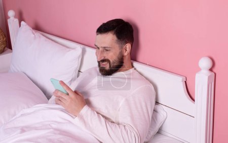 Foto de Joven barbudo en pijama usando teléfono móvil en la cama. - Imagen libre de derechos