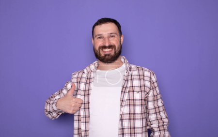 Photo for Young handsome bearded man standing over isolated purple background doing happy thumbs up gesture. Approving expression looking at the camera showing success. - Royalty Free Image