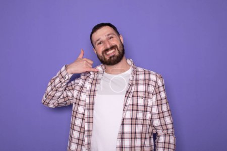 Téléchargez les photos : Jeune homme avec barbe faisant geste de téléphone sur fond violet - en image libre de droit