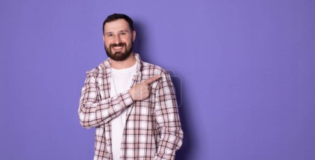 Téléchargez les photos : Beau barbu tendance élégant confiant positif homme joyeux pointant sur l'espace de copie vierge, sur fond violet pastel - en image libre de droit