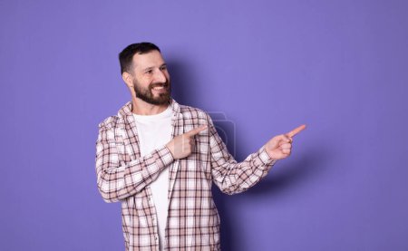 Foto de Guapo barbudo de moda elegante confiado hombre alegre positivo apuntando en espacio de copia en blanco, sobre fondo violeta pastel púrpura - Imagen libre de derechos