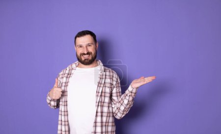 Foto de Guapo barbudo de moda elegante confiado hombre alegre positivo apuntando en espacio de copia en blanco, sobre fondo violeta pastel púrpura - Imagen libre de derechos