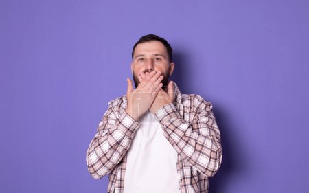 Téléchargez les photos : Jeune homme fermant la bouche avec les mains et regardant la caméra sur fond violet - en image libre de droit