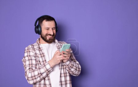 Foto de Retrato de un joven barbudo sonriente con auriculares usando un teléfono inteligente sobre fondo púrpura. - Imagen libre de derechos