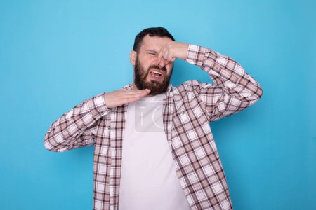 Photo for Caucasian bearded man isolated over blue background smelling something stinky and disgusting, intolerable smell, holding breath with fingers on nose. Bad smells concept. - Royalty Free Image