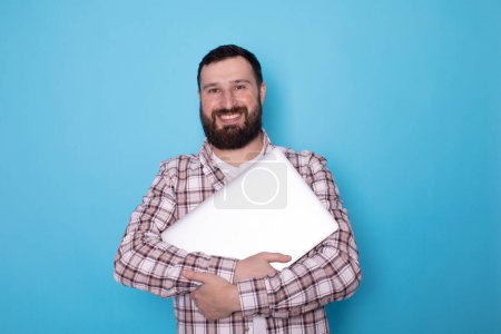 Téléchargez les photos : Sourire jeune homme barbu tenant ordinateur portable sur fond bleu - en image libre de droit