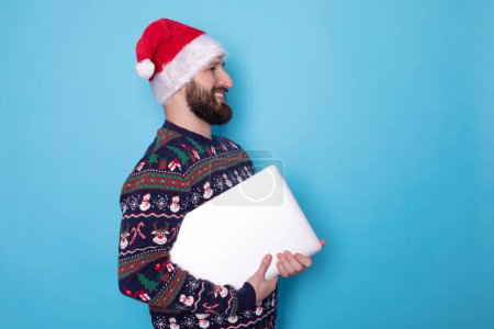Photo for Bearded man in knitted sweater and red Christmas Santa Claus hat hold in hands closed laptop pc computer isolated on blue background. Happy New Year 2024 celebration. - Royalty Free Image