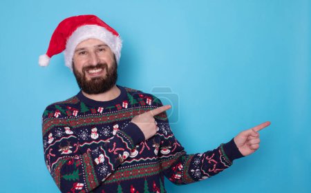 Téléchargez les photos : Joyeux jeune homme barbu en pull de Noël et chapeau de Père Noël pointant quelque chose sur fond bleu clair. Espace pour le texte. - en image libre de droit