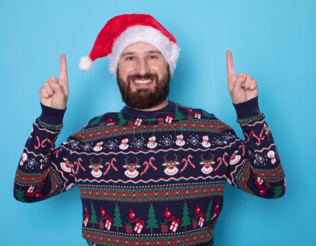 Téléchargez les photos : Joyeux jeune homme barbu en pull de Noël et chapeau de Père Noël pointant quelque chose sur fond bleu clair. Espace pour le texte. - en image libre de droit