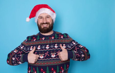 Photo for Portrait of handsome happy bearded man in santa hat and christmas sweater showing thumbs up on blue background - Royalty Free Image