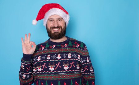 Photo for Portrait of handsome happy bearded man in santa hat and christmas sweater showing ok sign on blue background - Royalty Free Image