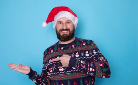 Photo for Happy young bearded man in Christmas sweater and Santa hat pointing at something on light blue background. Space for text. - Royalty Free Image