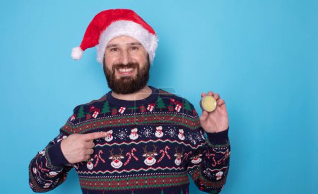 Photo for Handsome bearded man in santa hat and christmas sweater holding beard balm on blue background - Royalty Free Image