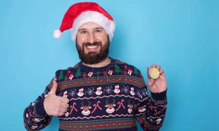 Téléchargez les photos : Bel homme barbu en chapeau de Père Noël et pull de Noël tenant baume barbe sur fond bleu - en image libre de droit