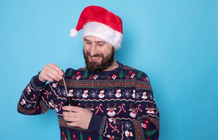 Photo for Bearded man in christmas sweater and santa hat holding pipette with beard oil. - Royalty Free Image