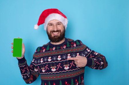 Téléchargez les photos : Joyeux homme barbu porter pull de Noël tricoté posant tenant téléphone portable avec espace de travail écran blanc isolé sur fond bleu. Bonne année 2024 concept de vacances. - en image libre de droit